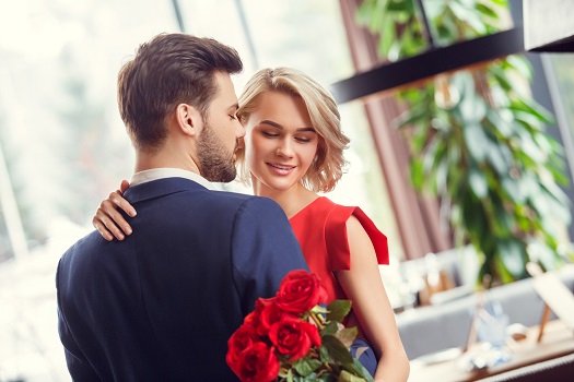 Young man and woman on date in restaurant dancing sensual together holding bouquet of red roses smiling passionate